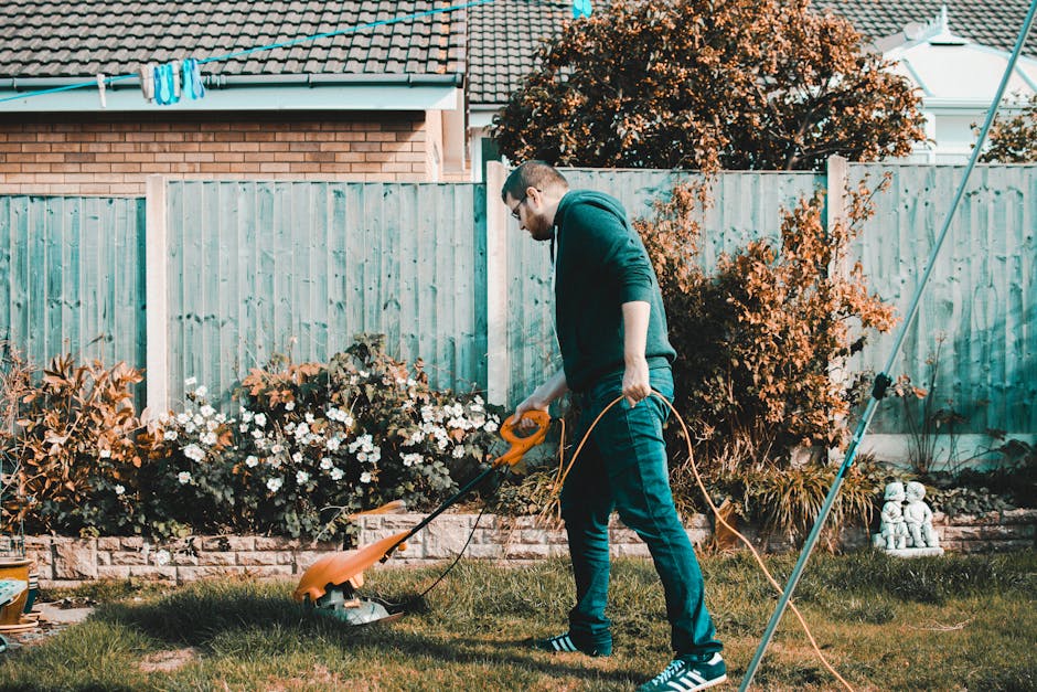 A gardener wearing a dark jacket, hat, and gloves is trimming a dense, green hedge with long-handled pruning shears. The hedge appears well-maintained, with lush foliage and light pink flowers visible