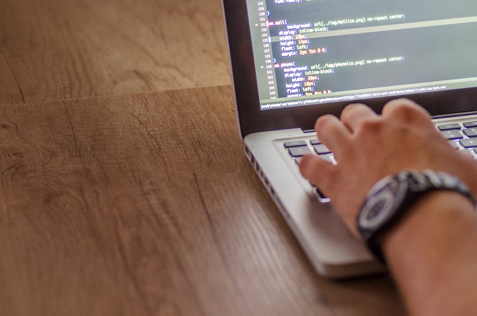 A silver laptop displaying lines of code on its screen, placed on a wooden table in a cozy indoor setting with warm lighting. The background features blurred shelves and furniture, suggesting a home o